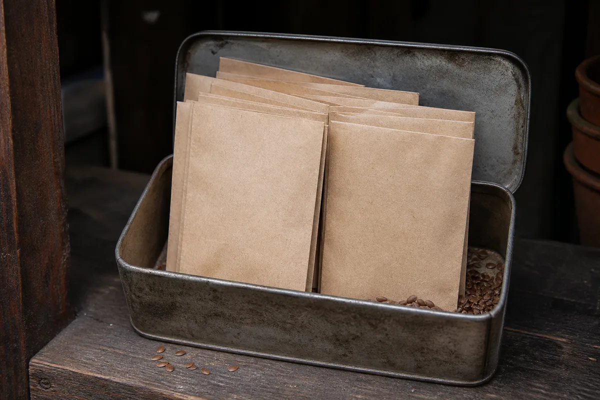 Sachets de graines en papier kraft rangés dans une boîte métallique fermée, dans une réserve sombre et fraîche
